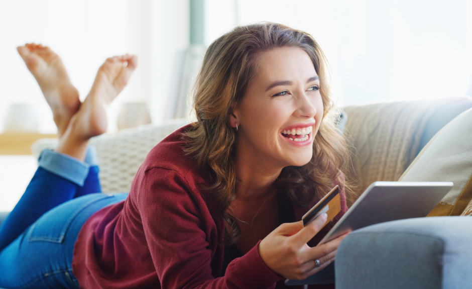 Frau liegt lachend auf der Couch und hat eine Kreditkarte, sowie ein Tablet in der Hand.