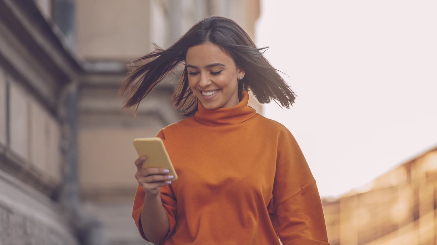 Frau mit orangem Pullover schaut im gehen auf ihr Handy und lächelt.