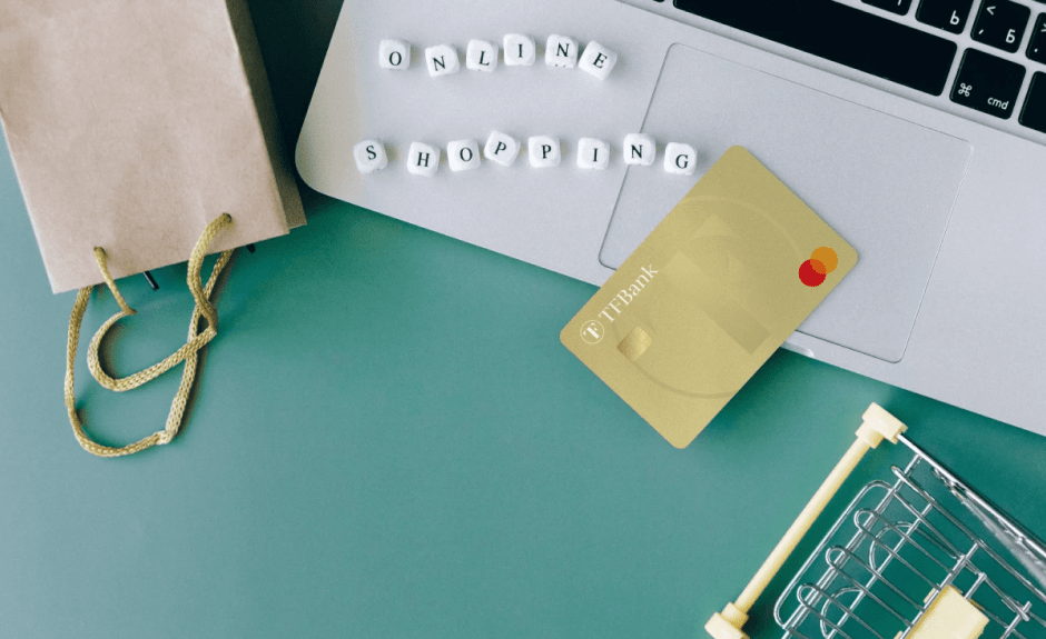 TF Mastercard Gold liegt auf Laptop. Daneben liegen kleine Würfel mit Buchstaben zusammengesetzt als "Online Shopping". Außerdem liegt dort eine kleine Tüte und eiin kleiner modelhafter Einkaufkorb.