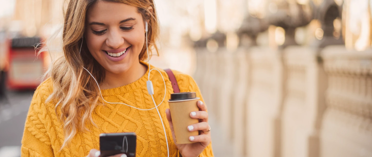 Frau läuft lächelnd  mit einem Cafe-to-go in der Hand und schaut auf ihr Handy. Neben ihr ist ein Smartphone abgebildet und zeigt die Funktion der Click to Pay Registierung.