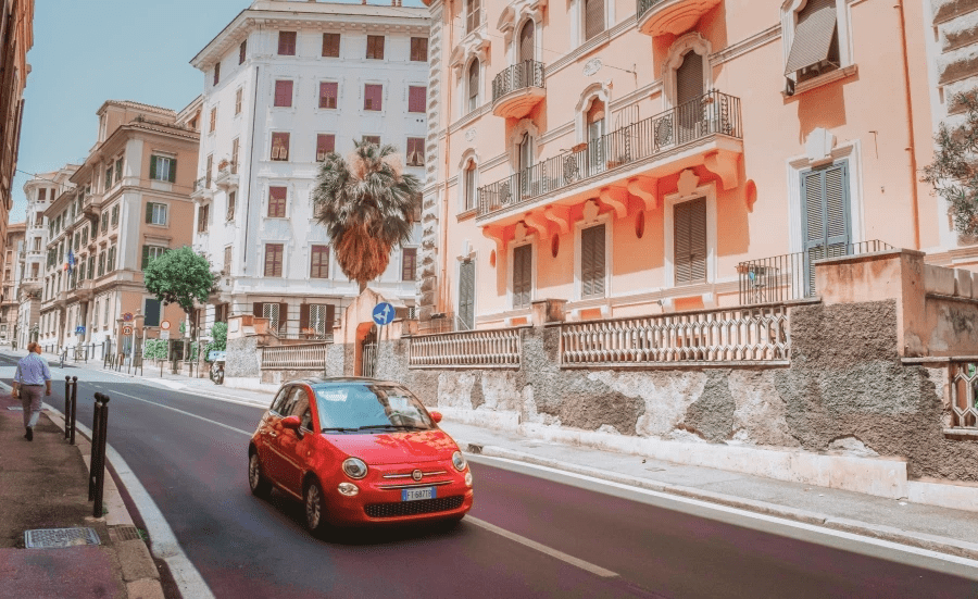 Auf einer Straße fährt ein rotes Auto. Im Hintergrund sind Häuser im südländischem Stil zu sehen.