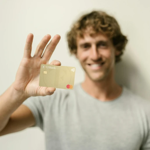 Ein Mann steht lächelnd vor einer Wand und hält die TF Mastercard Gold in die Kamera.